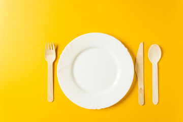 Tableware from natural materials - bamboo wooden fork, spoon and knife with white plate isolated on a yellow background.