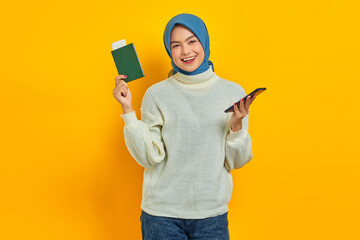 Cheerful beautiful Asian woman in white sweater holding mobile phone and passport isolated over yellow background. Summer Trip