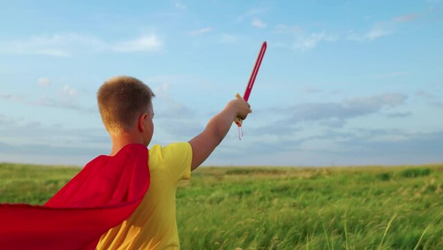 Child waves toy sword, childhood dreams. Child boy plays superhero. Child Game, Imagination. Boy, child in red cloak with sword raised in his hand up sword, on field, depicting medieval knight.