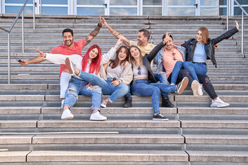 Fototapeta premium Group of cheerful multi-ethnic young men and women in casual outfits, smiling happily and looking at the camera as they sit on the stairs and hug each other after their successful studies.