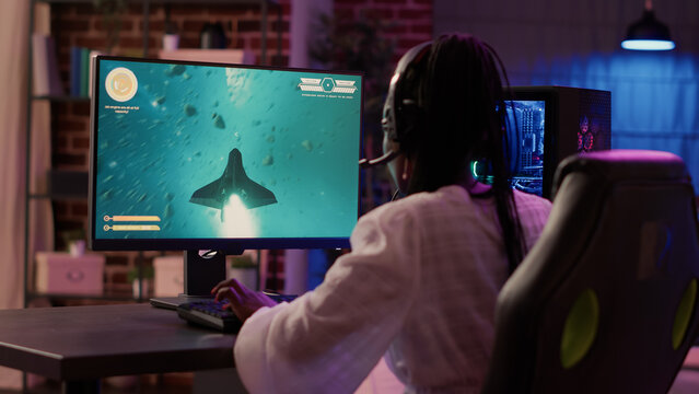 Over shoulder view of african american gamer girl playing space shooter simulation using pc gaming setup enjoying free time at home. Woman streaming online action game multiplayer competition.
