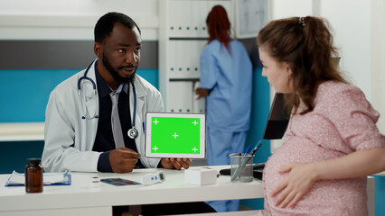 Male specialist holding tablet with horizontal greenscreen on display in cabinet. Doctor and pregnant woman looking at isolated chroma key background with blank mockup template. Tripod shot.