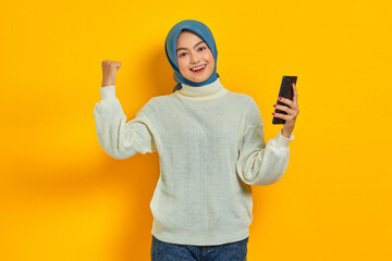 Smiling young asian woman in white sweater and hijab using mobile phone, doing winner gesture isolated over yellow background. People islam religious concept