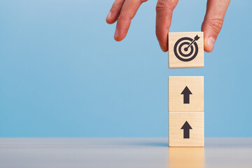 Hand putting stack of wooden blocks with arrow and target icons on a blue background copy space. Business development strategy, advancement and goal concept.