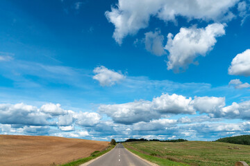 An asphalt road section in a rural area passing by agricultural fields against a background of blue sky and white fluffy clouds on a warm sunny day.