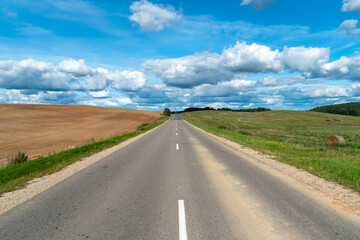 Fototapeta premium An asphalt road section in a rural area passing by agricultural fields against a background of blue sky and white fluffy clouds on a warm sunny day.