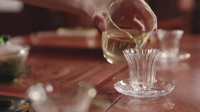 Chinese woman pours freshly prepared green tea into a glass cup on traditional dark wood tea table. Chinese tea ceremony.