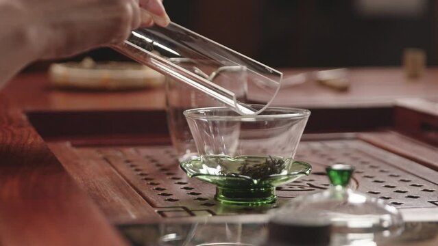 Asian woman puts green tea leaves from glass tray into Chinese gaiwan bowl on traditional wooden tea table. Close up.