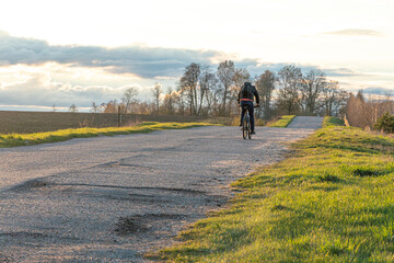 Obraz premium A cyclist rides on an asphalt road in the countryside during sunset past beautiful fields and flower meadows. A receding single cyclist on an empty road.