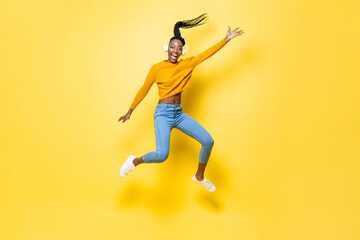 Fototapeta na wymiar Positive enthusiastic young African American woman wearing headphones listening to music and jumping with hand up in yellow isolated studio background