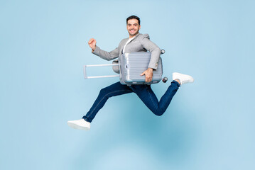 Excited fun Caucasian tourist man with baggage, ready to travel, jumping in studio light blue color...