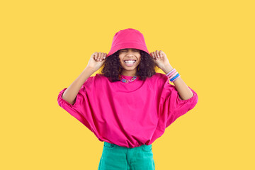 Happy joyful kid in trendy casual outfit puts pink bucket hat on her head. Cheerful beautiful Black girl wearing loose fuchsia top and green pants having fun and laughing. Children's fashion concept - obrazy, fototapety, plakaty