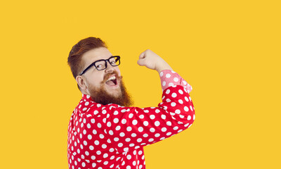 Funny stylish bearded chubby man with smile on face shows showing biceps on yellow background. Joyful crazy redhead millennial man in glasses and red shirt in white peas shows his power. Web banner. - obrazy, fototapety, plakaty