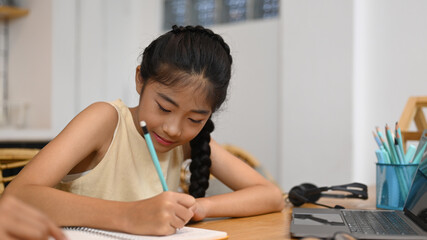 Fototapeta na wymiar Asian school girl distance learning online, doing homework while sitting at kitchen table