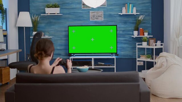 Gamer girl playing console video games with wireless controller on green screen tv relaxing on couch in living room. Young woman enjoying free time at home doing online gaming on chroma key display.