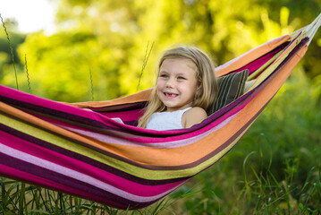 A preschooler girl sits in a bright striped hammock in a park in nature on a warm sunny day