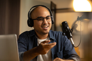 Young asian man host streaming podcast with condenser microphone work on laptop at small broadcast...