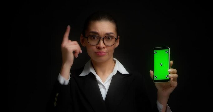 Business Woman With Glasses And Office Clothes Holds A Horizontal Smartphone With A Green Screen For Copy Space, Looks At Him, Shrugs, I Don't Know. Black Background. Chroma Key Mock-up