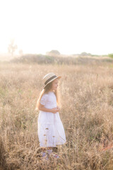 A cute little girl with long blond curly hair in a white summer dress and a straw boater hat in a field in the countryside in summer at sunset. Nature and Ecolife
