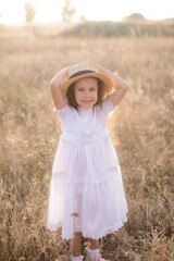 A cute little girl with long blond curly hair in a white summer dress and a straw boater hat in a field in the countryside in summer at sunset. Nature and Ecolife