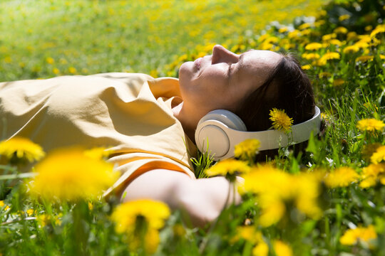 Audio Healing. Meditation. Slow Life. Enjoying The Little Things. Dreaming Of Spring. Middle Aged Woman Wearing Headphones Listens To Music Lying On The Grass With Dandelions On A Spring Day. Burnout 