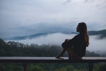 Portrait image of a woman sitting on wooden balcony with a beautiful mountains and nature view on foggy day