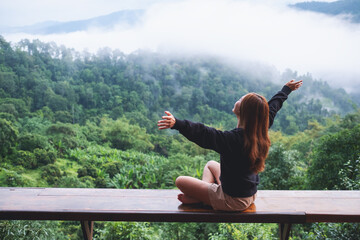 A young woman with arms raised sitting on wooden balcony with a beautiful mountains view on foggy...