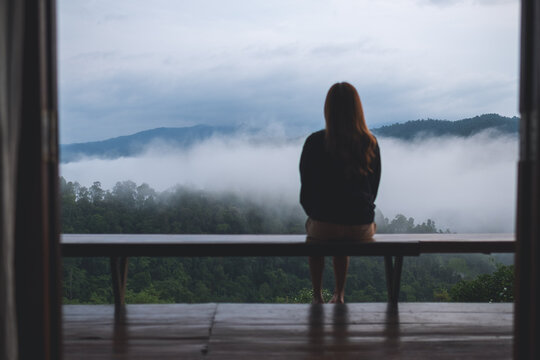 Rear view image of a young woman sitting alone on wooden balcony and looking at a beautiful foggy mountain and nature view