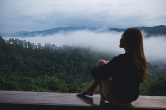 Portrait image of a woman sitting on wooden balcony with a beautiful mountains and nature view on foggy day