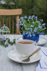 A white cup of tea and flowers in a small blue vase on an old wooden table in the garden.