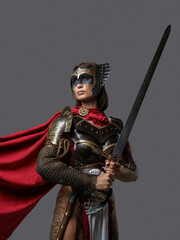 Fototapeta premium Shot of female barbarian holding sword dressed in steel armor with helmet against grey background.