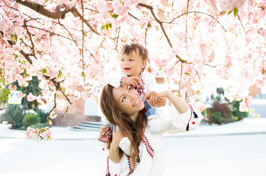 Little Boy On Hands Of Mother. Woman Playing With Child Outside In Blooming Spring Garden. Portrait Of Family Of Two People. Spring Landscape Background.