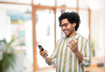 technology, communication and people concept - happy smiling young man in glasses with smartphone over office background - obrazy, fototapety, plakaty