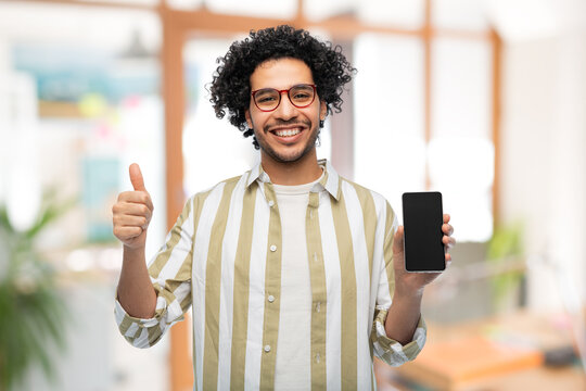 technology and people concept - smiling man in glasses holding smartphone with blank screen and showing thumbs up gesture over office background