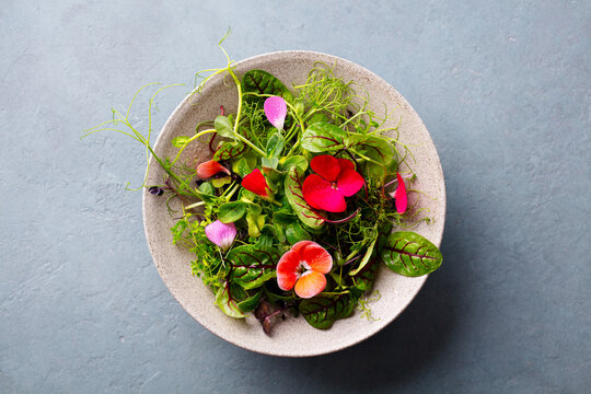 Green salad leaves, sprouts with edible flowers in bowl. Grey background. Close up. Top view.