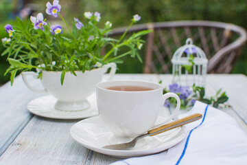 A white cup with tea and flowers in a small white vase on an old wooden table in the garden.