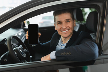 Handsome young car salesman sitting in auto salon, showing blank mobile phone with mockup for...