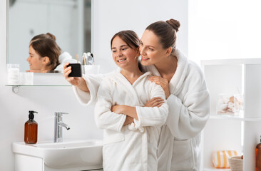 beauty, hygiene, morning and people concept - happy smiling mother and daughter taking selfie with smartphone at bathroom