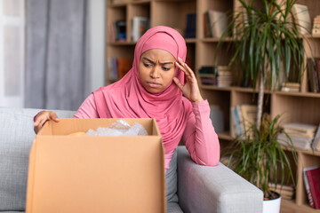 Wrong item. Upset black muslim lady looking at open parcel with despair, unpacking box while...