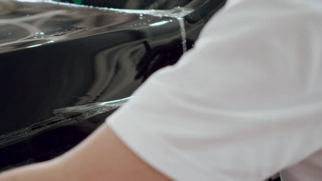 A professional employee of a car wash or detaling studio cuts off a piece of film from the hood of a black car. Close-up of the hand of the washer glues a protective film on the car.