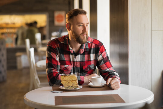 Young bearded man drinking coffee with a cake in the morning in a cafe, a businessman starting his working day with breakfast in a restaurant