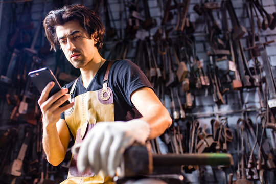 Authentic blacksmith man wear leather apron in dark indoors studio looking at phone screen
