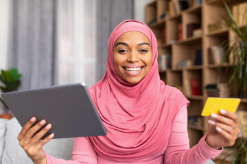 Happy black muslim lady shopping from home, using digital tablet and credit card, buying things in web store