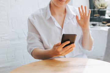 A woman in a white shirt waves and communicates via video call. Online communication and training by phone. Freelance marketer works through the phone in a cafe