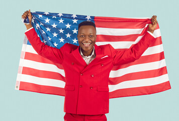 Happy millennial dark skinned man raises american national flag on pastel light blue background. Joyful man in red suit with large fabric flag of America smiling at camera. Concept of patriotism.