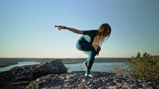 Gorgeous woman practicing in Garudasana - eagle pose, yoga on high mountain above water. Free and wild. Flexibility workout at nature background. Strong lady strengthening neck and back, equilibrium