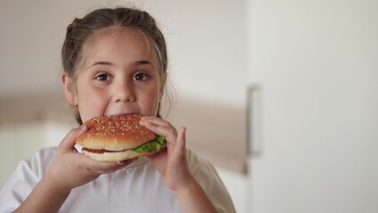 little girl eating a hamburger. unhealthy fast food proper nutrition concept. child greedily with pleasure bites a big burger in the kitchen at home lifestyle. kid eats fast food close-up