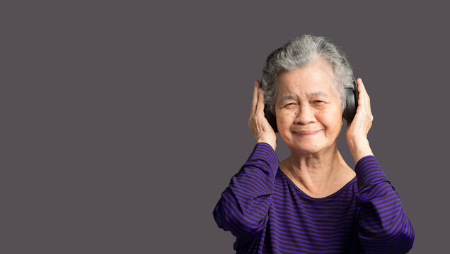 Cheerful elderly Asian woman wearing wireless headphones with a smile and looking at the camera while standing on a gray background
