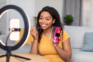 Beauty blog concept. Happy black makeup artist recording cosmetics product review, showing her lipstick palette - obrazy, fototapety, plakaty