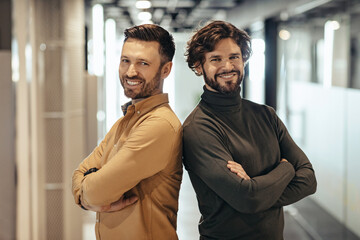 Portrait of two happy businessmen standing with crossed arms, smiling at camera in modern company hall